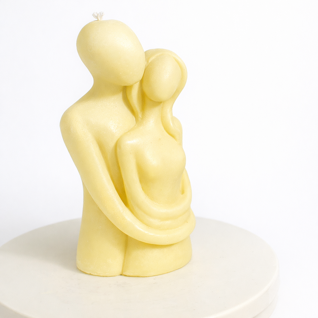 Vela escultórica minimalista en color amarillo suave con figura de pareja abrazada, elaborada en cera natural premium, diseño contemporáneo romántico sobre base circular, pieza decorativa elegante ideal para regalo de aniversario o decoración moderna.