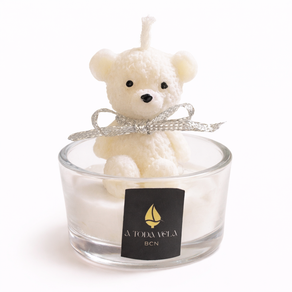 Vela decorativa artesanal con osito de cera color marfil en vaso de cristal transparente, diseño infantil de lujo con lazo plateado, fotografía de estudio minimalista para regalo premium y decoración delicada.