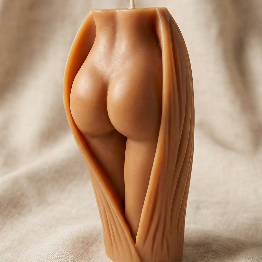 Vela decorativa escultural con forma de glúteos y piernas femeninas emergiendo de una envoltura orgánica texturizada. La vela tiene un tono de piel mestiza cálido y está situada sobre un fondo de tela beige suave.