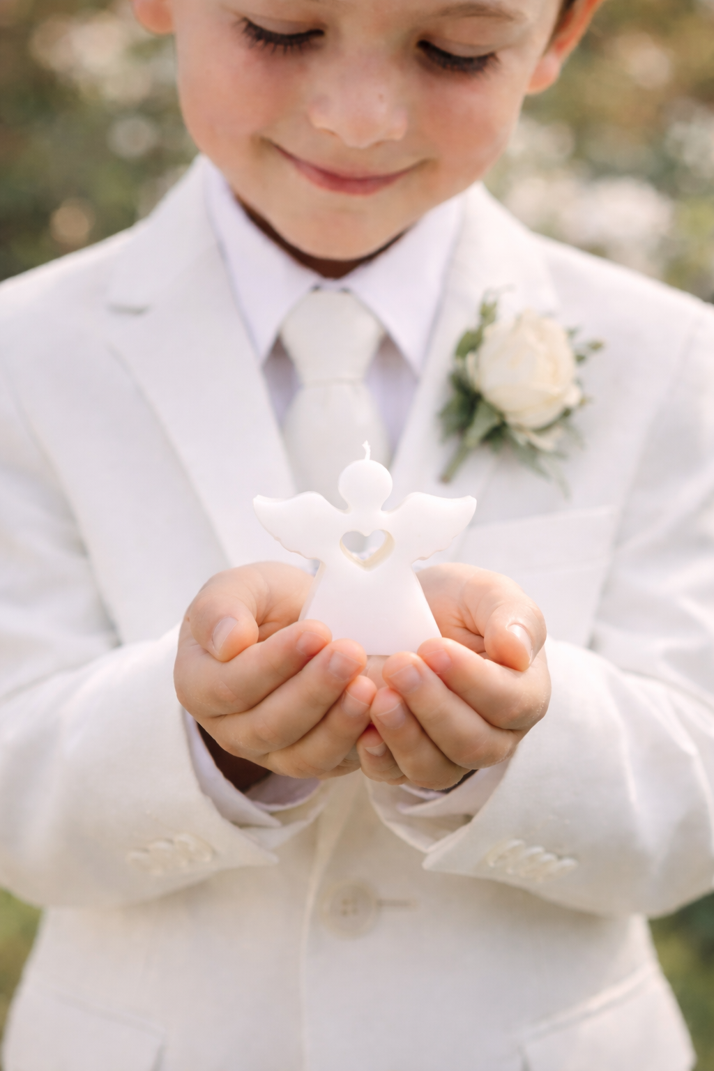 Niño vestido de blanco sosteniendo vela artesanal en forma de ángel blanco durante celebración de comunión o evento religioso elegante al aire libre.