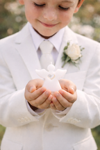 Niño vestido de blanco sosteniendo vela artesanal en forma de ángel blanco durante celebración de comunión o evento religioso elegante al aire libre.
