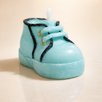 Vela de diseño en forma de zapatito de bebé azul pastel, elaborada en cera artesanal con textura textil y cordones decorativos, fotografía de producto premium con iluminación suave.