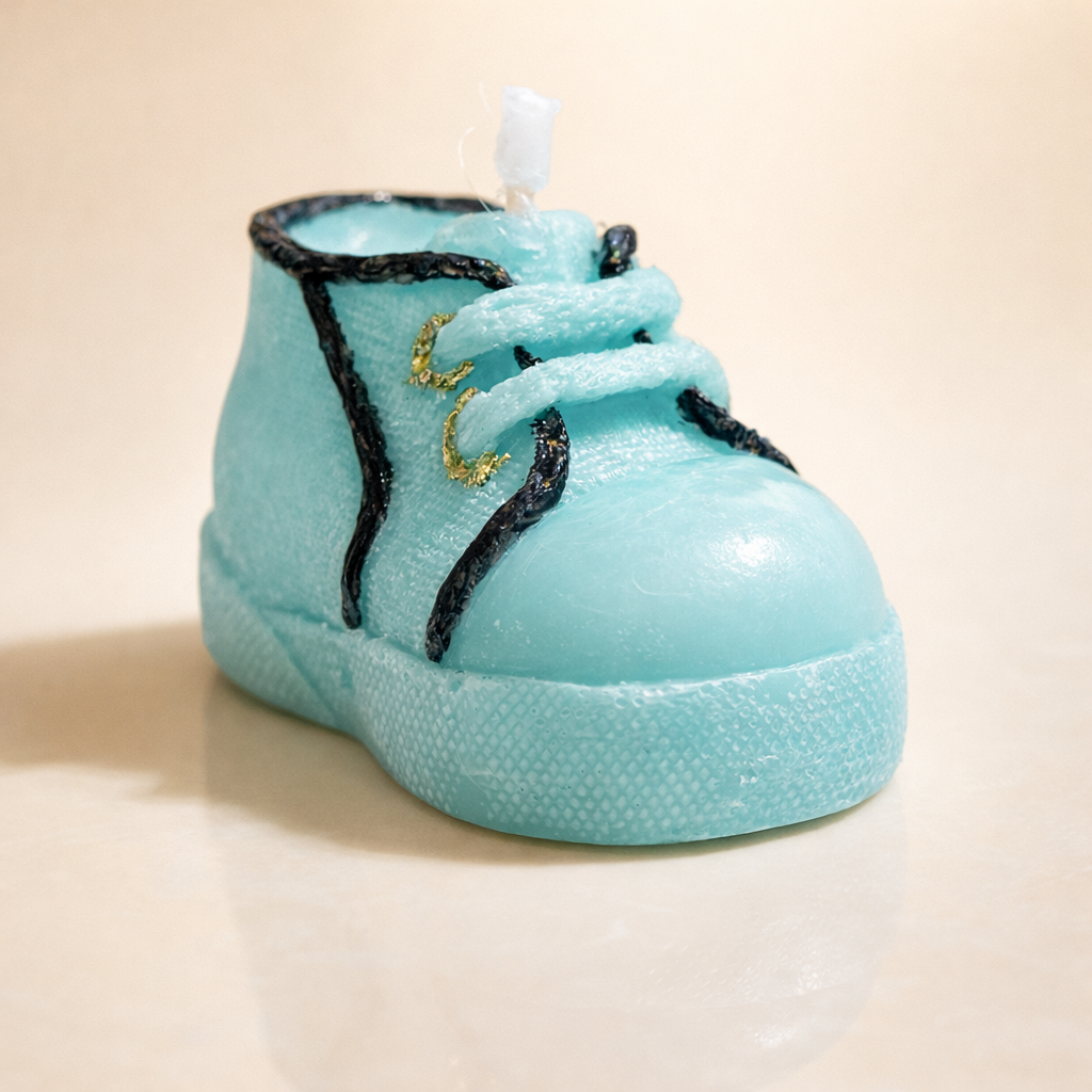 Vela de diseño en forma de zapatito de bebé azul pastel, elaborada en cera artesanal con textura textil y cordones decorativos, fotografía de producto premium con iluminación suave.
