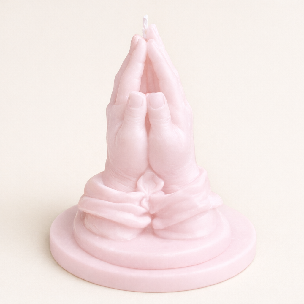 Vela artística en forma de manos rezando color rosa empolvado, realizada en cera natural con acabado delicado, diseño escultórico elegante sobre base circular, objeto decorativo exclusivo para interiores sofisticados.