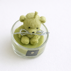 Vela infantil de lujo con osito de cera verde natural en vaso de cristal, textura suave y acabado artesanal, fotografía de producto elegante para decoración y regalo exclusivo.