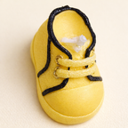 Vista superior de vela artesanal en forma de zapatito de bebé color amarillo suave, realizada en cera natural con acabados detallados, fotografía de producto elegante con estética editorial contemporánea.