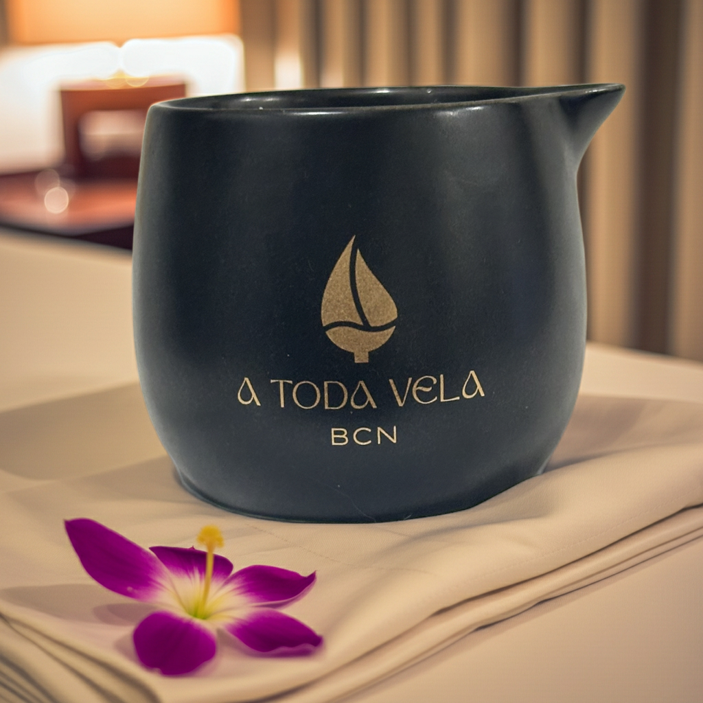 Vela Masaje Yuuyake Lujo, en vaso cerámico negro con logo dorado "A Toda Vela BCN", sobre una servilleta y una flor orquídea morada.