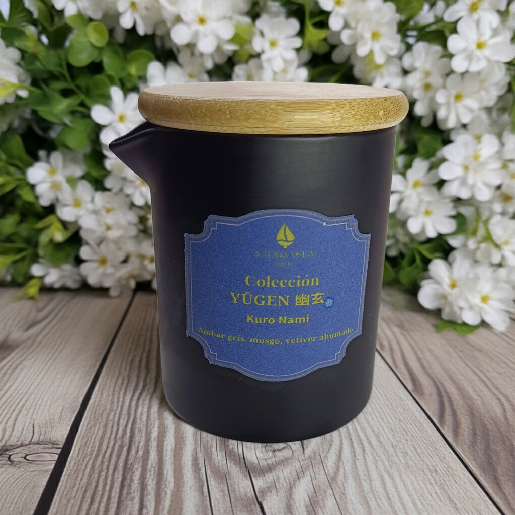 Vela Masaje Kuro Nami Lujo, en recipiente cerámico negro con tapa de bambú y etiqueta de Colección YŪGEN, sobre fondo de flores blancas.