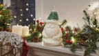 Vela Papá Noel Gorro Árbol, figura tallada de Santa Claus con gorro de pino verde y barba blanca, sobre repisa navideña con luces y guirnaldas.