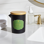 Vela Masaje Satori no Kō en vaso cerámico negro con tapa de bambú, sobre un lavabo de mármol blanco para ritual de baño.